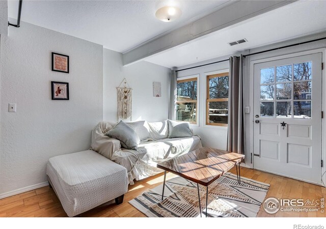 living area featuring a textured wall, visible vents, baseboards, light wood finished floors, and beamed ceiling