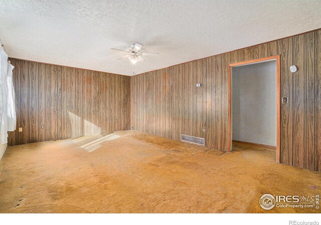 carpeted empty room with wood walls, ceiling fan, visible vents, and a textured ceiling