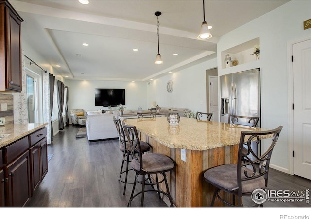 kitchen with dark wood-style flooring, hanging light fixtures, light stone countertops, stainless steel fridge, and a kitchen breakfast bar