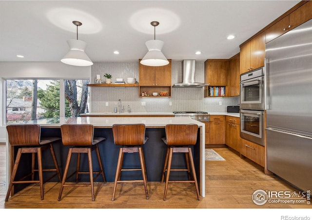 kitchen with stainless steel appliances, a sink, light countertops, wall chimney range hood, and brown cabinets