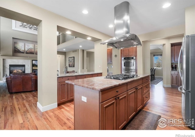 kitchen with light stone counters, island range hood, stainless steel appliances, light wood-type flooring, and a glass covered fireplace