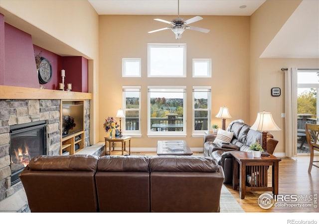 living area featuring baseboards, light wood-style flooring, a high ceiling, ceiling fan, and a stone fireplace