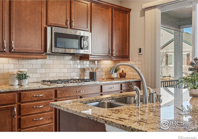 kitchen with brown cabinets, stainless steel appliances, decorative backsplash, a sink, and light stone countertops