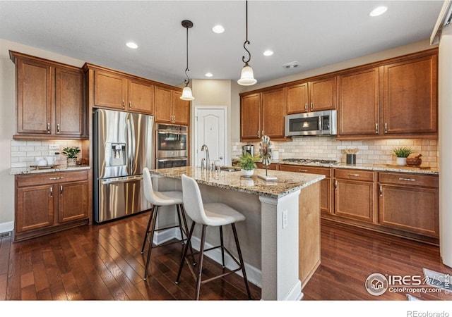 kitchen with dark wood-style floors, appliances with stainless steel finishes, brown cabinets, light stone counters, and a kitchen bar