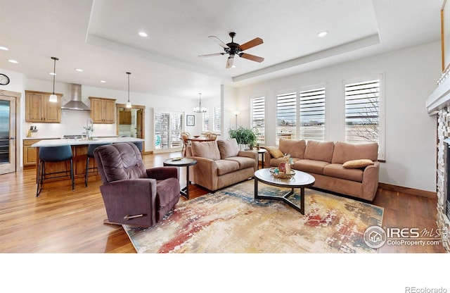 living area with a fireplace, recessed lighting, a raised ceiling, light wood-style floors, and ceiling fan with notable chandelier