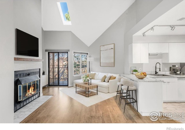 living room featuring a skylight, light wood finished floors, a fireplace with flush hearth, track lighting, and high vaulted ceiling