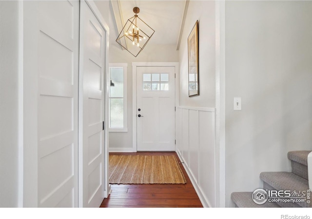 doorway to outside with dark wood-style floors, stairs, a notable chandelier, and wainscoting