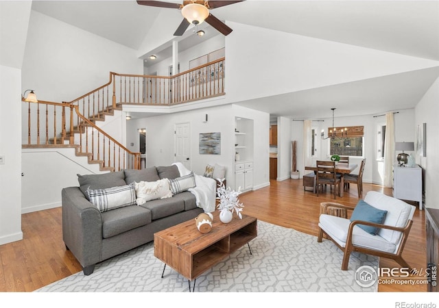 living area featuring stairway, wood finished floors, baseboards, high vaulted ceiling, and ceiling fan with notable chandelier