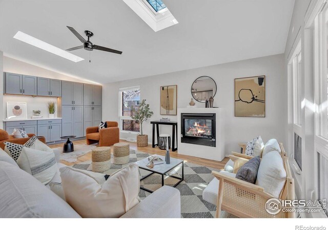 living room with a ceiling fan, a glass covered fireplace, light wood-type flooring, and vaulted ceiling with skylight