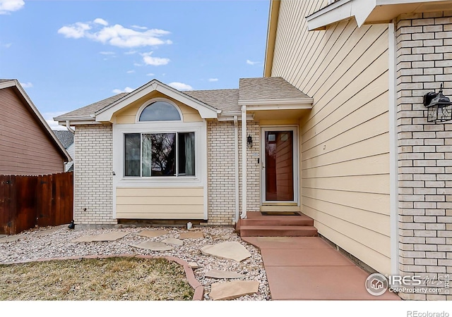 doorway to property featuring brick siding, a shingled roof, and fence