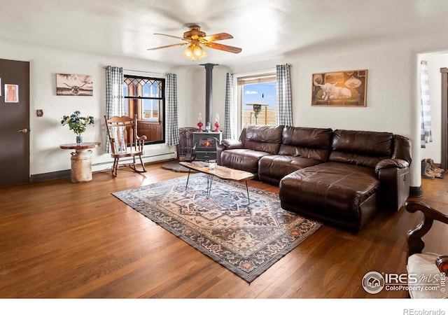 living room with a baseboard radiator, wood finished floors, a wood stove, and a ceiling fan