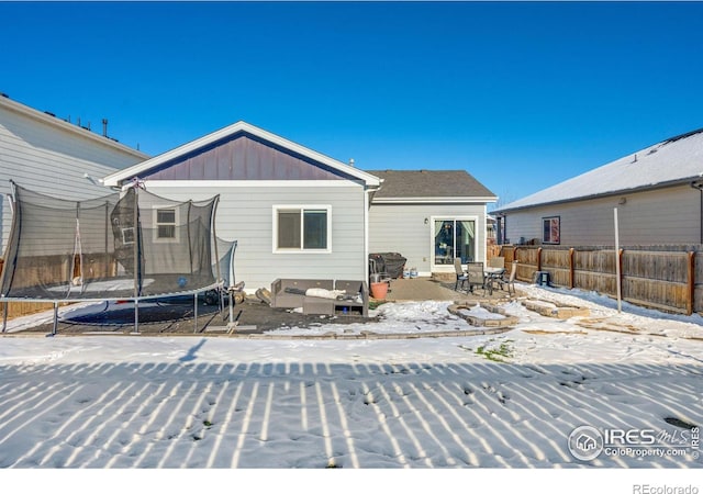 snow covered back of property with a trampoline, fence, board and batten siding, and a patio