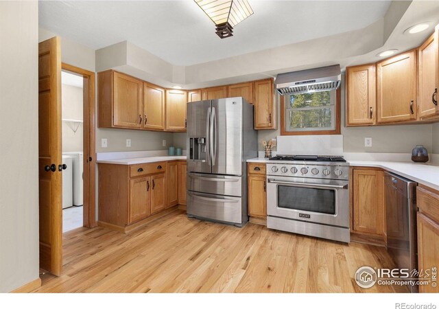 kitchen featuring light wood finished floors, appliances with stainless steel finishes, brown cabinets, light countertops, and wall chimney range hood