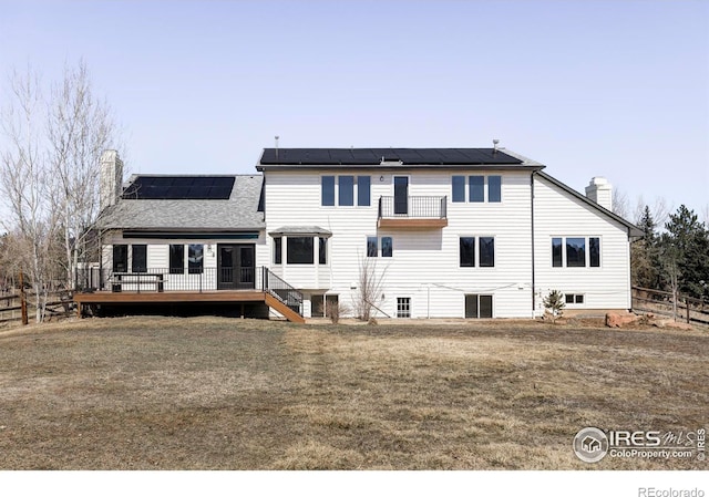 back of property with a deck, fence, a lawn, roof mounted solar panels, and a chimney