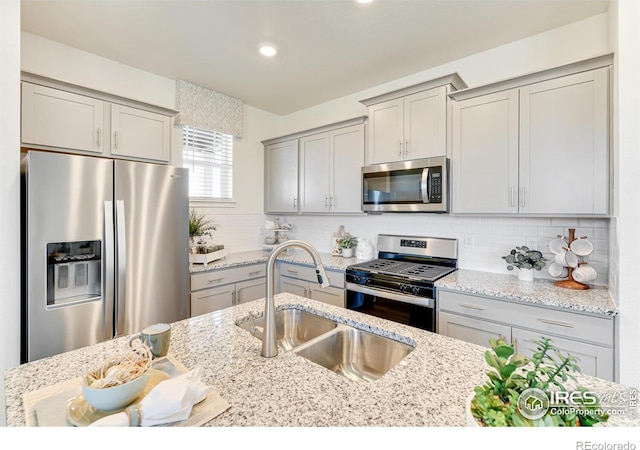 kitchen with light stone counters, gray cabinetry, a sink, appliances with stainless steel finishes, and tasteful backsplash