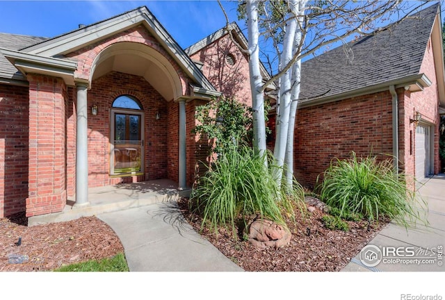 view of exterior entry featuring a shingled roof, brick siding, and an attached garage