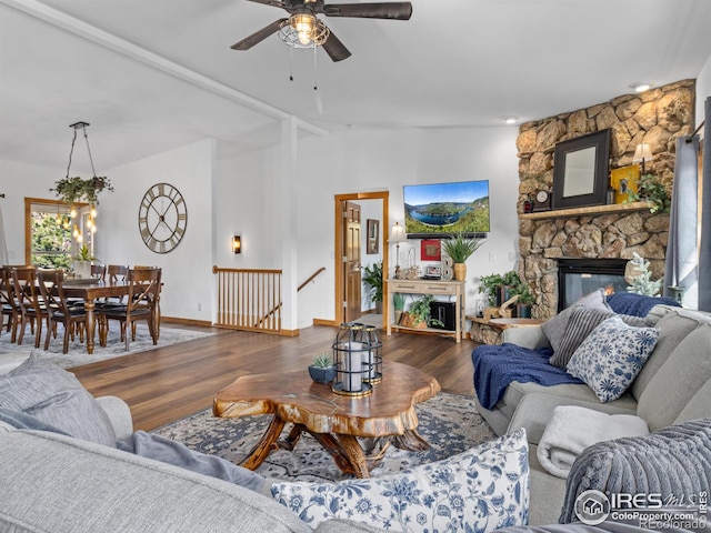 living room with ceiling fan, a stone fireplace, wood finished floors, baseboards, and vaulted ceiling