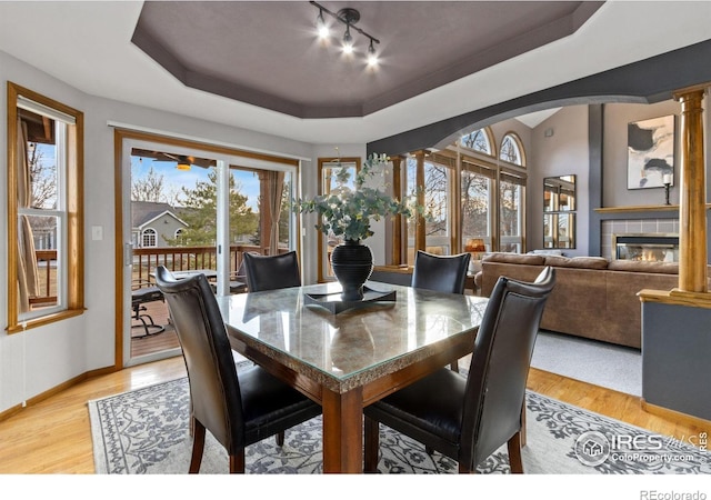 dining room with ornate columns, a tray ceiling, light wood finished floors, and a tiled fireplace