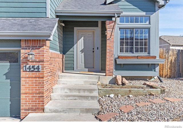 entrance to property featuring a garage, brick siding, fence, and roof with shingles