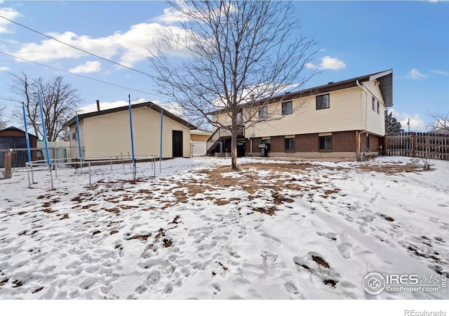 snow covered rear of property with a trampoline, brick siding, a storage unit, fence, and an outdoor structure