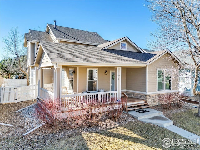 view of front of house with stone siding, a shingled roof, fence, and covered porch