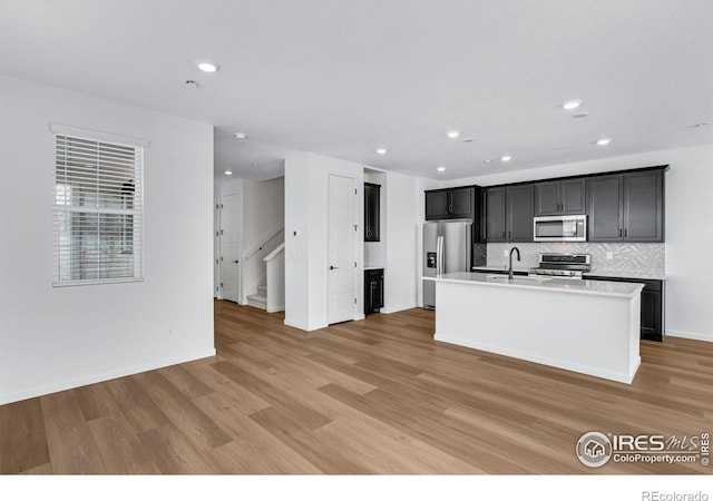 kitchen featuring decorative backsplash, appliances with stainless steel finishes, a kitchen island with sink, light countertops, and light wood-type flooring