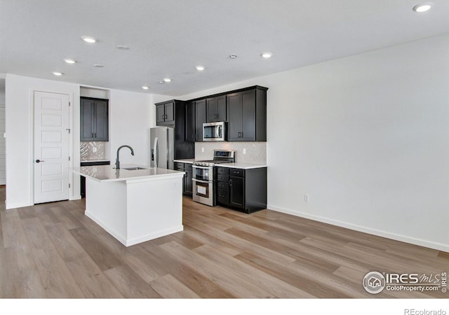 kitchen with a kitchen island with sink, light wood-style flooring, a sink, light countertops, and appliances with stainless steel finishes