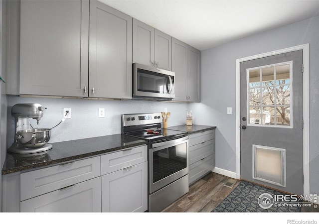 kitchen with gray cabinetry, baseboards, appliances with stainless steel finishes, dark stone counters, and dark wood finished floors