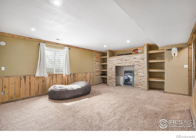 carpeted living room featuring built in shelves, wainscoting, wood walls, a stone fireplace, and a textured ceiling