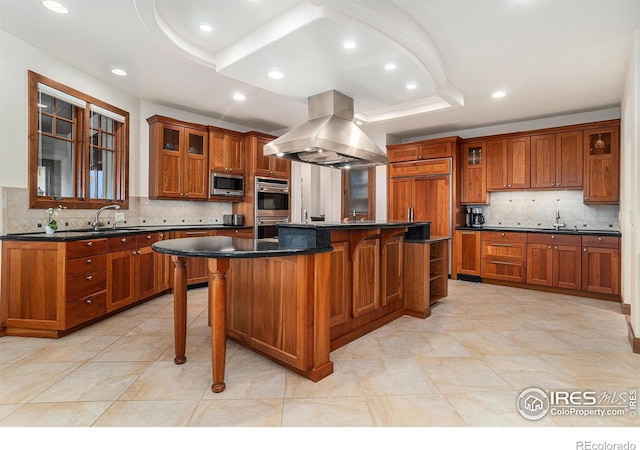 kitchen featuring built in appliances, brown cabinets, a tray ceiling, dark countertops, and island exhaust hood