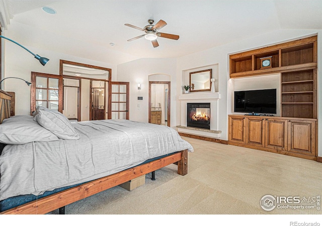 bedroom featuring arched walkways, light colored carpet, a glass covered fireplace, vaulted ceiling, and ensuite bath