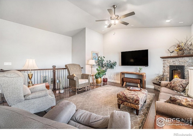 living area featuring ceiling fan, vaulted ceiling, a stone fireplace, wood finished floors, and baseboards