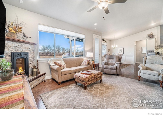 living area featuring a stone fireplace, vaulted ceiling, light wood-style flooring, and ceiling fan with notable chandelier