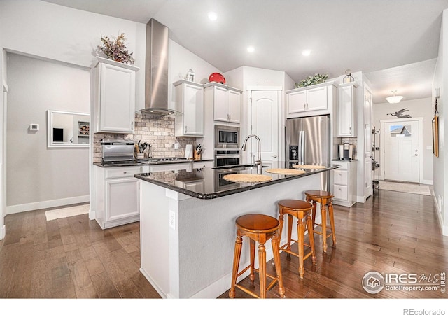 kitchen with dark countertops, wall chimney exhaust hood, appliances with stainless steel finishes, a kitchen island with sink, and a sink