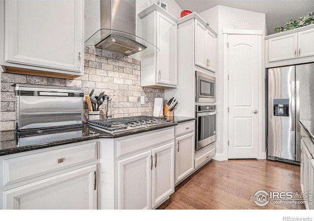 kitchen with wall chimney exhaust hood, light wood-style flooring, appliances with stainless steel finishes, white cabinetry, and backsplash