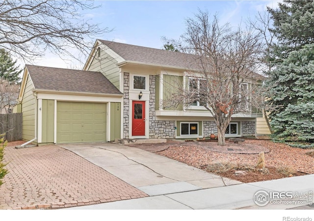 tri-level home featuring a shingled roof, concrete driveway, fence, a garage, and stone siding