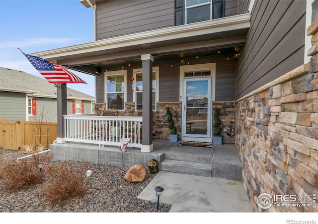 view of exterior entry with covered porch, stone siding, and fence