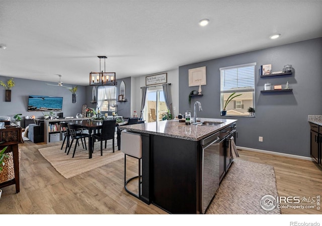 kitchen with light wood-style flooring, stainless steel dishwasher, stone countertops, a sink, and an island with sink