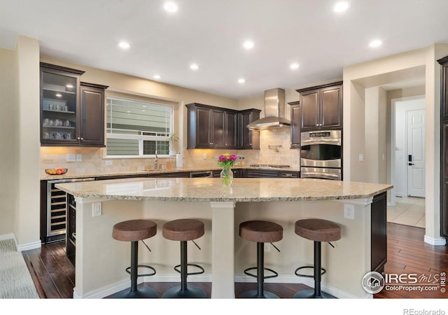 kitchen featuring light stone counters, a sink, dark brown cabinets, appliances with stainless steel finishes, and wall chimney exhaust hood