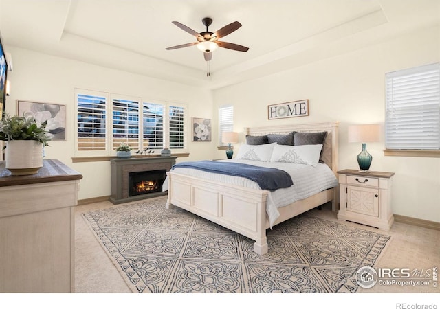 bedroom with a ceiling fan, a warm lit fireplace, a raised ceiling, and baseboards