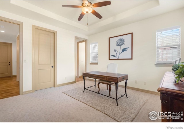 home office featuring ceiling fan, baseboards, a raised ceiling, and light colored carpet