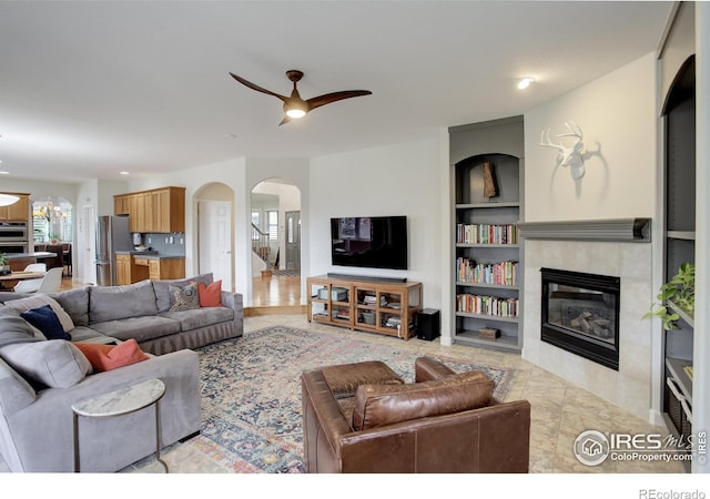 living area featuring built in features, arched walkways, recessed lighting, a tile fireplace, and ceiling fan with notable chandelier