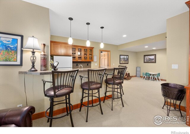 kitchen with brown cabinetry, freestanding refrigerator, glass insert cabinets, light carpet, and a kitchen bar