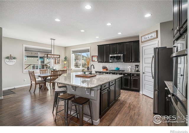 kitchen with a center island with sink, appliances with stainless steel finishes, a breakfast bar, dark wood-style flooring, and backsplash