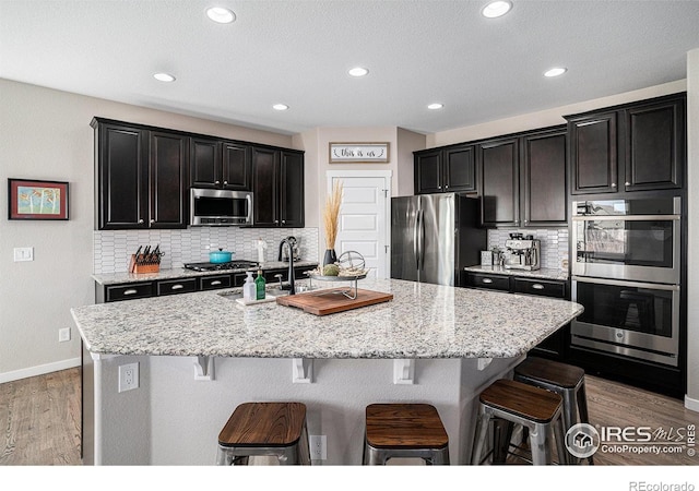 kitchen with a sink, a kitchen breakfast bar, appliances with stainless steel finishes, light stone countertops, and an island with sink