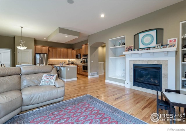 living room featuring visible vents, arched walkways, a tile fireplace, light wood-type flooring, and built in shelves