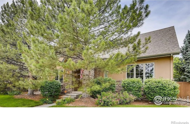 view of property hidden behind natural elements featuring stone siding, roof with shingles, and stucco siding