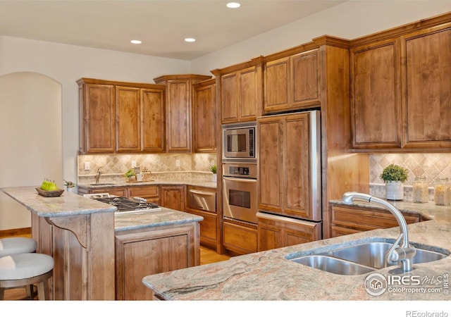 kitchen featuring light stone countertops, brown cabinetry, built in appliances, and a sink