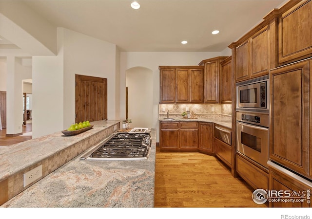 kitchen featuring backsplash, light stone countertops, light wood-style flooring, brown cabinetry, and stainless steel appliances