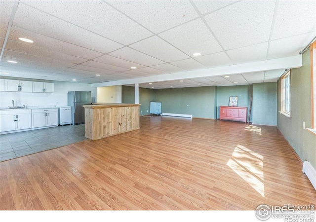 unfurnished living room with light wood finished floors, bar area, a baseboard heating unit, a drop ceiling, and a sink